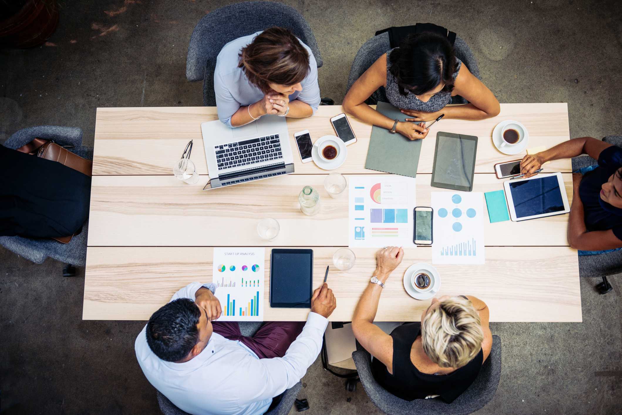 Top down view of people sitting at a table collaborating on a project. There are laptops and sheets of paper with graphs strewn about the table.